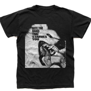May include: Black t-shirt featuring a grayscale image of a snarling dog's face with the text "BITE THE HAND THAT STARVES YOU" in a bold, sans-serif font. The graphic is centered on the shirt, creating a striking visual contrast.
