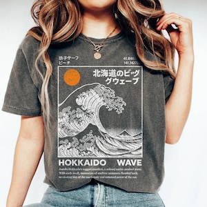 May include: Charcoal grey t-shirt with a white and black graphic depicting a large wave, a rising sun, and Japanese text. The shirt also features the text "HOKKAIDO WAVE" and further text below the graphic. A relaxed fit tee.