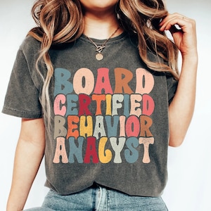 Peut inclure: Un t-shirt gris foncé avec le texte "BOARD CERTIFIED BEHAVIOR ANALYST" dans une police arc-en-ciel rétro.
