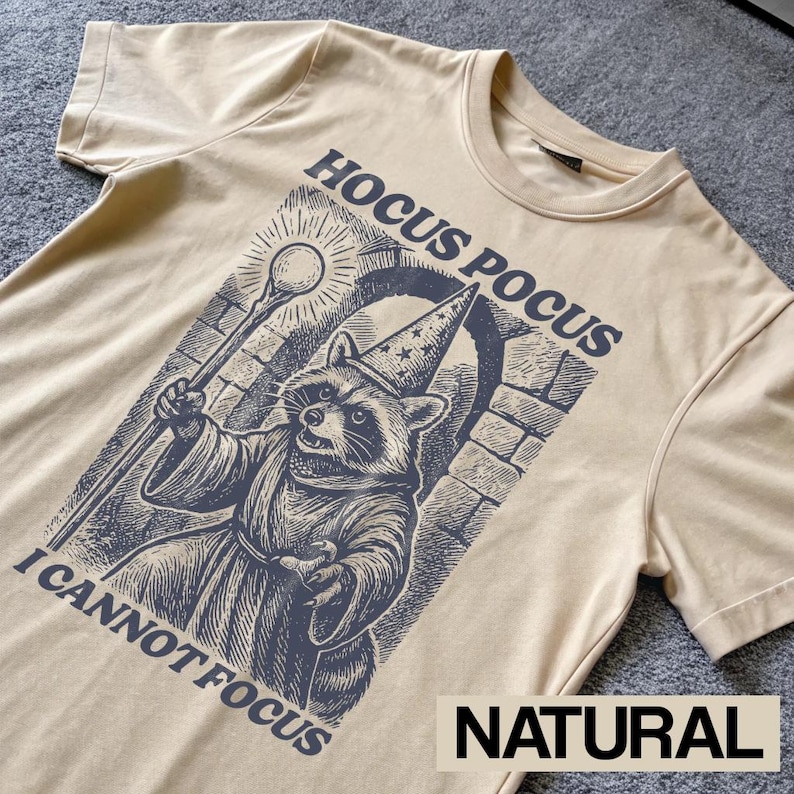 May include: A natural-colored t-shirt featuring a raccoon dressed as a wizard, holding a staff. The graphic includes the text "Hocus Pocus I Cannot Focus" in dark blue, with the word "NATURAL" at the bottom.