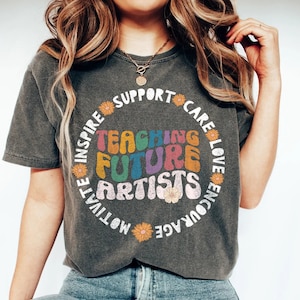 May include: A dark gray t-shirt with a colorful graphic that says "Teaching Future Artists" in a retro font. The graphic is surrounded by a circle of flowers and the words "Inspire, Motivate, Encourage, Love, Care, Support".