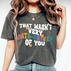 Könnte beinhalten: Dunkelgraues T-Shirt mit dem Text "THAT WASN'T VERY DATA-DRIVEN OF YOU" in Weiß und bunten Buchstaben. Das Shirt hat einen Vintage-Look, leicht verwaschen. Kleine Blumenmotive sind um den Text platziert.