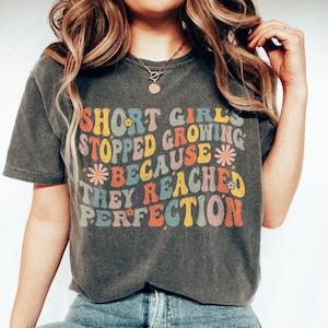 Könnte beinhalten: Ein graues T-Shirt mit einer Retro-Grafik, auf der "Kleine Mädchen haben aufgehört zu wachsen, weil sie die Perfektion erreicht haben" steht. Die Grafik ist in bunten, regenbogenartigen Buchstaben mit Blumen.