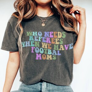 Puede incluir: Una camiseta gris con el texto "¿Quién necesita árbitros cuando tenemos mamás de fútbol?" en colores arcoíris.