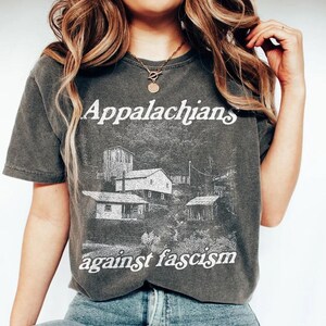May include: Dark gray t-shirt with the words "Appalachians against fascism" in white. The shirt features a black and white illustration of buildings and trees. The shirt is a short-sleeved crew neck.