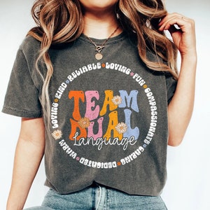 Könnte beinhalten: Ein graues T-Shirt mit einer bunten Grafik, auf der "Team Dual Language" steht, umgeben von einem Kreis aus Wörtern, die positive Eigenschaften beschreiben. Die Wörter sind "liebend, freundlich, zuverlässig, mitfühlend, fürsorglich, engagiert, warmherzig, lustig, inspirierend".