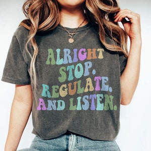 Puede incluir: Una camiseta gris oscuro con el texto "Alright Stop. Regulate. And Listen." en letras de colores de estilo retro.