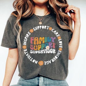 Può includere: Una maglietta grigio scuro con un design grafico colorato che dice "Family Support Supervisor" circondato da un cerchio di fiori e parole come "ispirare", "motivare", "incoraggiare", "prendersi cura", "amare" e "sostenere".