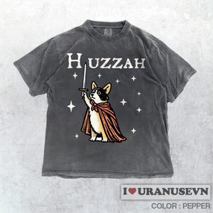 K&ouml;nnte beinhalten: Dunkelgraues T-Shirt mit dem Wort "Huzzah" &uuml;ber einem Cartoon-Corgi-Hund, der ein Schwert h&auml;lt und einen Umhang tr&auml;gt. Das Design enth&auml;lt Sterne. Der Text "I love URANUSEVN" und "COLOR: PEPPER" befindet sich unten.