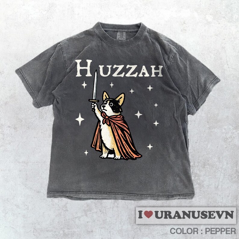 K&ouml;nnte beinhalten: Dunkelgraues T-Shirt mit dem Wort "Huzzah" &uuml;ber einem Cartoon-Corgi-Hund, der ein Schwert h&auml;lt und einen Umhang tr&auml;gt. Das Design enth&auml;lt Sterne. Der Text "I love URANUSEVN" und "COLOR: PEPPER" befindet sich unten.
