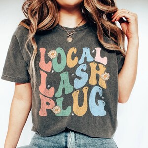 Pode incluir: Uma camiseta cinza escura com um gráfico de estilo retrô que diz "LOCAL LASH PLUG" em letras coloridas com detalhes de margaridas.