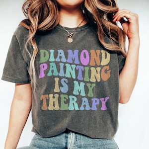 May include: A woman wearing a gray t-shirt with the text "DIAMOND PAINTING IS MY THERAPY" in rainbow colors. The shirt is a relaxed fit and has a distressed look.