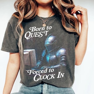 May include: Dark gray t-shirt with the text "Born to QUEST Forced to CLOCK IN." The graphic depicts a knight in shining armor at a computer. The shirt has a vintage, worn look.