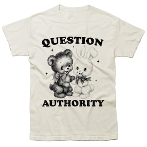 May include: Cream-colored t-shirt featuring a black and white graphic of a teddy bear and a bunny holding hands. The text "QUESTION AUTHORITY" is printed above and below the graphic in a bold, black font. The design includes small star accents.