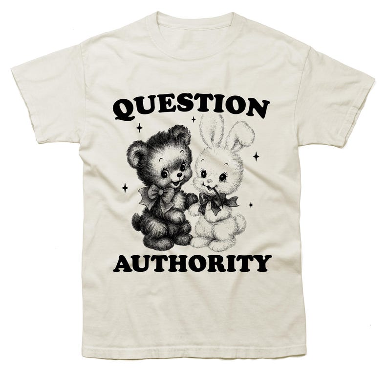 May include: Cream-colored t-shirt featuring a black and white graphic of a teddy bear and a bunny holding hands. The text "QUESTION AUTHORITY" is printed above and below the graphic in a bold, black font. The design includes small star accents.