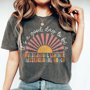May include: A dark gray t-shirt with a retro sun graphic and the text "It's a good day to be a Multilingual Learner Instructional Coach".