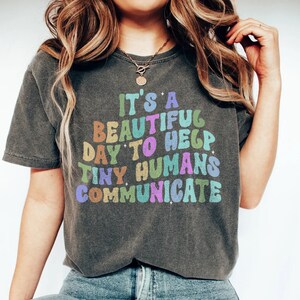 Może przedstawiać: Szary t-shirt z napisem "It's a beautiful day to help tiny humans communicate" w kolorach tęczy.