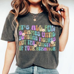 May include: A dark grey t-shirt with a colourful graphic that reads "It's all fun & games until someone figures out the function of your behaviour".