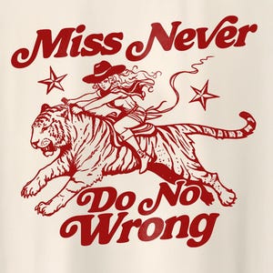 Könnte beinhalten: Cremefarbenes Grafik-T-Shirt mit einer roten Illustration einer Frau, die einen Tiger reitet. Der Text "Miss Never Do No Wrong" ist oberhalb und unterhalb des Bildes gewölbt, mit Sternen auf beiden Seiten. Das Design hat eine Vintage- und Retro-Ästhetik.