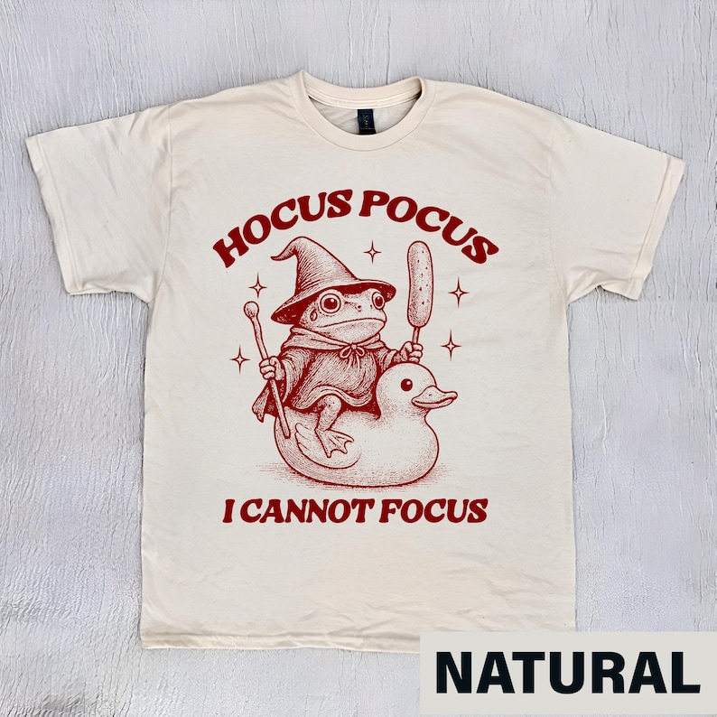Puede incluir: Camiseta de color natural con un gr&aacute;fico rojo. El gr&aacute;fico presenta una rana con sombrero y capa de mago montada en un pato, con las palabras "Hocus Pocus I Cannot Focus". La camiseta es de material suave.