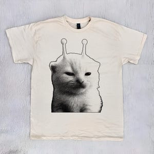 Op de afbeelding: Crèmekleurig T-shirt met een zwart-wit illustratie van een kat met antennes. De kat heeft een chagrijnige uitdrukking. Het shirt is gemaakt van een zacht materiaal en heeft een ronde hals.