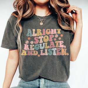 May include: A dark grey t-shirt with a retro-style graphic that reads "Alright Stop Regulate And Listen." The graphic is in colourful, wavy letters with small flower accents.