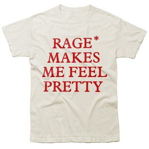 May include: Cream-coloured t-shirt featuring the red text "RAGE MAKES ME FEEL PRETTY". The short-sleeved top has a round neck and the text is printed in a bold font. The shirt is made of a soft material.