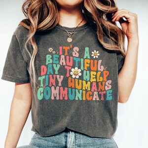 Może przedstawiać: Szary t-shirt z kolorowym nadrukiem, na którym widnieje napis "It's a beautiful day to help tiny humans communicate".