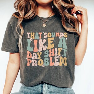 Könnte beinhalten: Ein dunkelgraues T-Shirt mit einer Retro-Grafik, auf der "That sounds like a day shift problem" steht. Die Grafik ist in bunten, abgerundeten Buchstaben mit kleinen Blumenakzenten gehalten.
