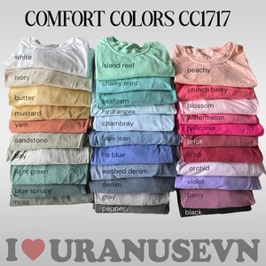 K&ouml;nnte beinhalten: Gestapelte, gefaltete T-Shirts in verschiedenen Farben, darunter Wei&szlig;, Elfenbein und Pfirsich. Das Bild zeigt den Text "COMFORT COLORS CC1717" und eine Liste von Farbbezeichnungen. Der untere Teil des Bildes enth&auml;lt den Text "I URANUSEVN".