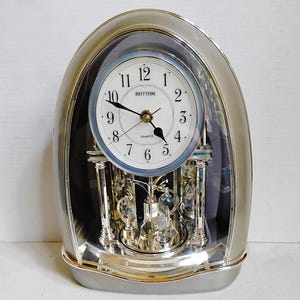 May include: An oval-shaped Rhythm quartz mantel clock with a silver-tone frame and a white face featuring black Arabic numerals. The clock includes a glass dome, internal silver pillars, and decorative crystal accents that rotate at the base.