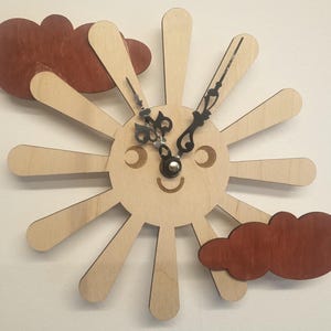 May include: A wooden wall clock shaped like a smiling sun with radiating beams. Two dark brown cloud cutouts are positioned on either side. The clock hands are black, and the face has a cheerful expression.