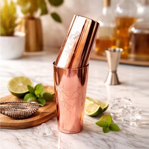 Op de afbeelding: Een koperkleurige cocktailshaker met geëtste bloemmotieven en de tekst "NBKdecor". De shaker is gedeeltelijk open. Limoenen, munt en ijsblokjes zijn rond de shaker gerangschikt, wat wijst op een cocktailthema.