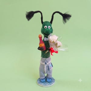 Op de afbeelding: Een groen vilten wezenfiguurtje met zwarte pijpenrager-antennes, gekleed in een lichtblauwe spijkerbroek en een kleine vlinderdas. Het figuurtje houdt een miniatuur rode fles en een klein boeket lichtroze rozen in doorzichtig plastic vast.