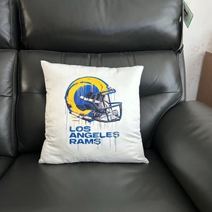 May include: White throw pillow featuring a blue and yellow Los Angeles Rams helmet graphic with paint splatters. The text "Los Angeles Rams" is printed below the helmet.
