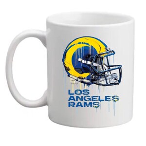 Peut inclure: Mug en céramique blanche avec un graphique de casque de football américain des Los Angeles Rams bleu et jaune. Le casque a un effet de peinture qui coule et le texte "Los Angeles Rams" est imprimé sous le casque.