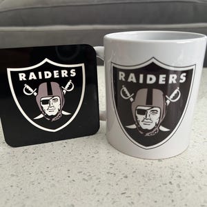 Op de afbeelding: Een witte keramische mok en een vierkante zwarte onderzetter, beide voorzien van het Raiders-logo. Het logo bestaat uit een schildvorm met de tekst RAIDERS boven een afbeelding van een persoon met een footballhelm, ooglapje en gekruiste zwaarden.