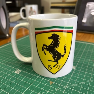 Può includere: Una tazza in ceramica bianca con un logo Ferrari nero e giallo. Il logo presenta un cavallo nero che si alza sulle zampe posteriori con le lettere "SF" sotto. La tazza ha una striscia verde, bianca e rossa intorno alla parte superiore.