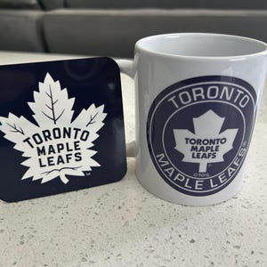 May include: A white ceramic mug and a square dark blue coaster, both featuring the Toronto Maple Leafs logo. The logo consists of a white maple leaf with the text Toronto Maple Leafs printed in dark blue. The mug also includes the NHL logo.