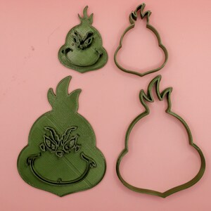 May include: Four green Grinch-shaped cookie cutters on a pink background. Two cutters are solid, showing the Grinch's face, while the other two are outlines. The Grinch's face is detailed with eyes, a nose, and a smiling mouth.