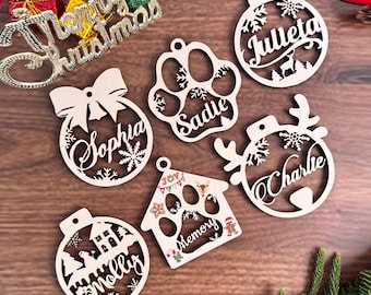 May include: Wooden Christmas ornaments in various shapes, including a paw print, a house, and round ornaments with names like Sophia, Charlie, and Molly. The ornaments are laser cut with snowflake and bow details. A "Merry Christmas" sign is also visible.