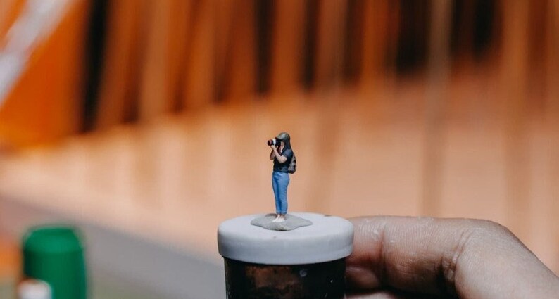 May include: A miniature figurine of a person holding a camera, standing on a small white platform. The figurine is wearing a blue shirt, blue jeans, and a black hat.