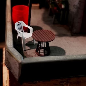 Puede incluir: Una silla de plástico blanca en miniatura y una pequeña mesa de madera marrón con una tapa rayada. La mesa y la silla están sobre una superficie gris.