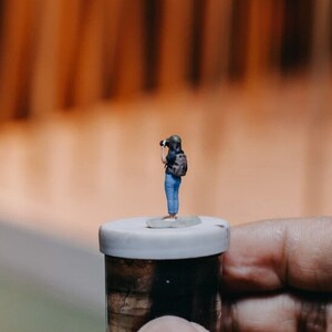 May include: A miniature figurine of a person holding a camera, standing on a small white lidded jar. The jar is made of glass and has a cork top.