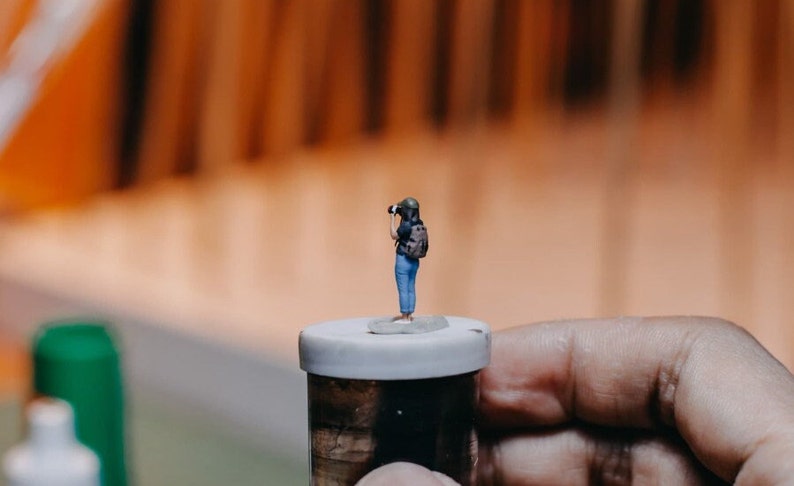 May include: A miniature figurine of a person holding a camera, standing on a small white lidded jar. The jar is made of glass and has a cork top.