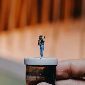 May include: A miniature figurine of a person wearing a blue shirt and a backpack, holding a camera, standing on top of a small, brown container with a white lid.