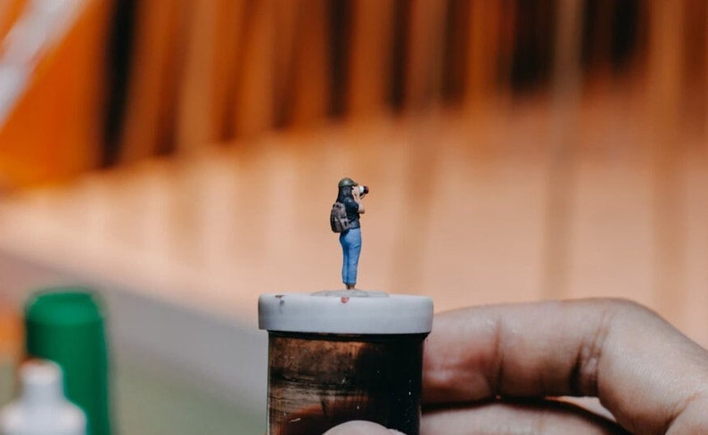 May include: A miniature figurine of a person wearing a blue shirt and a backpack, holding a camera, standing on top of a small, brown container with a white lid.