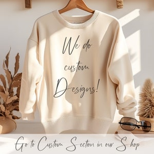 Pu&ograve; includere: Una felpa color crema con la scritta "We do custom Designs!" stampata sul davanti. Il testo &egrave; in un font scritto a mano. La felpa &egrave; appesa a un appendiabiti di legno. La scritta "Go to Custom Section in our Shop" &egrave; stampata sotto la felpa.