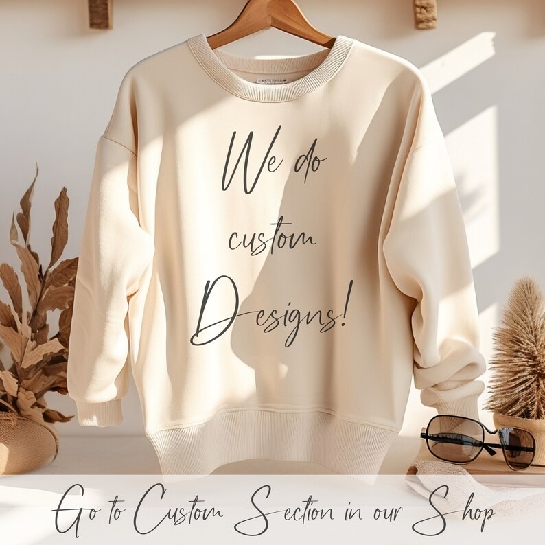 Pu&ograve; includere: Una felpa color crema con la scritta "We do custom Designs!" stampata sul davanti. Il testo &egrave; in un font scritto a mano. La felpa &egrave; appesa a un appendiabiti di legno. La scritta "Go to Custom Section in our Shop" &egrave; stampata sotto la felpa.