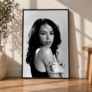 May include: A black and white portrait print in a thin black frame featuring a person with long dark hair looking over their shoulder. The person is wearing a dark top and a ring on their finger, captured in a high-contrast monochromatic style.